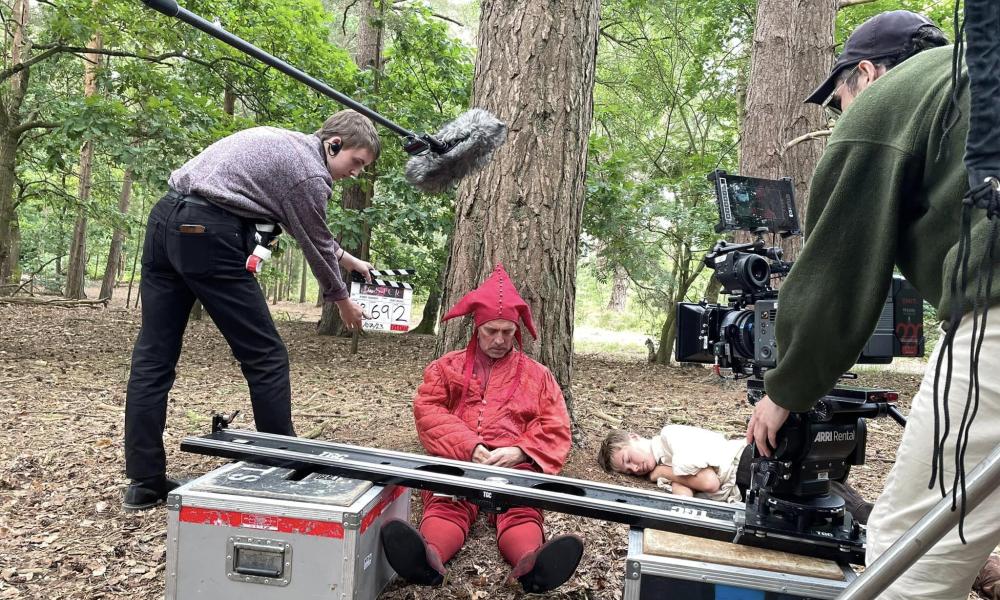 person dressed in red jester suit sat at foot of tree with camera and clapper board production team